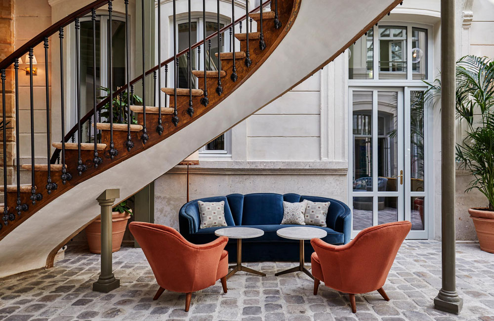 A blue sofa and two orange lounge chairs sit on cobbled stones underneath a spiral staircase
