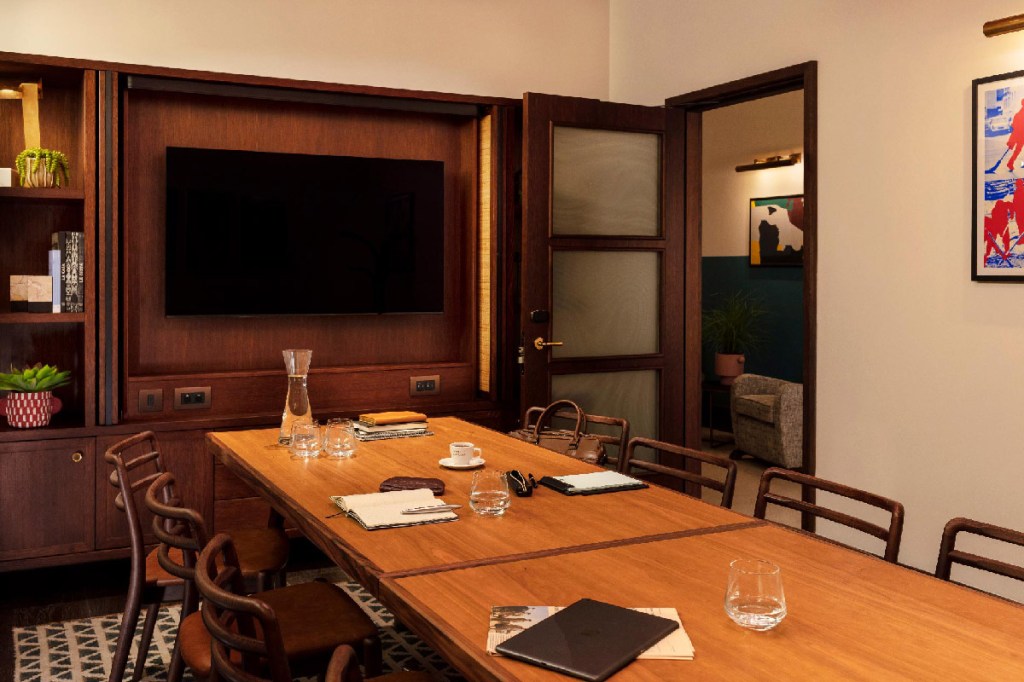 Meeting room with long table and 8 visible chairs, with large TV screen at the end of the table. Water, books, pencils and a laptop are on the table.