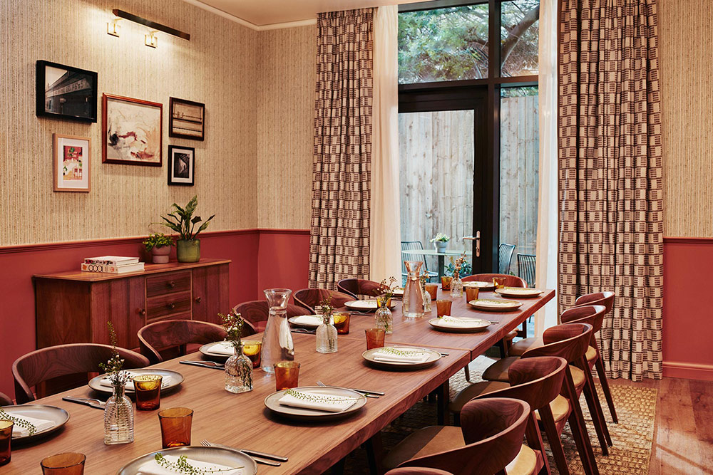 A long table arranged for a meal directs the eye to a large window with curtains open and seating outside.